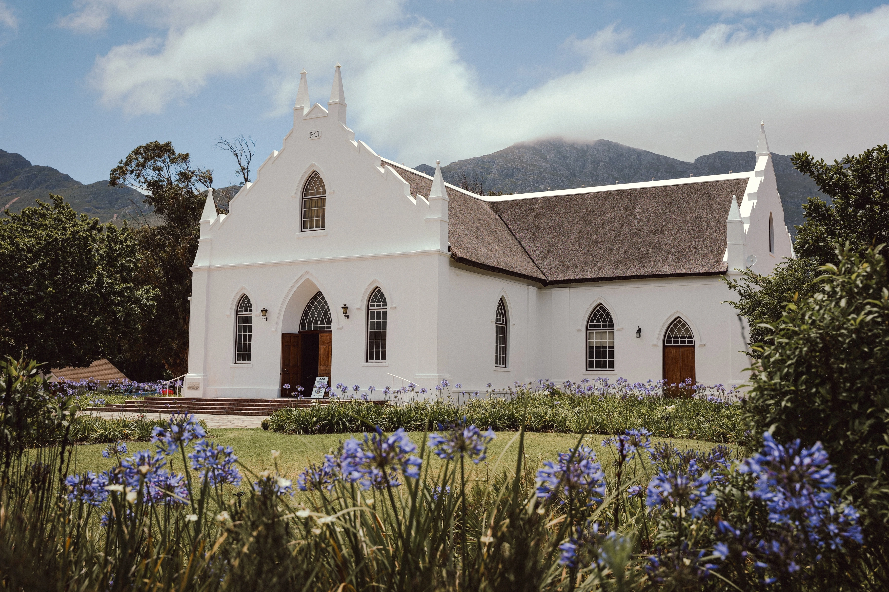 A white church with a thatched roof stands behind purple flowers, beneath a mountain ridge and cloud.