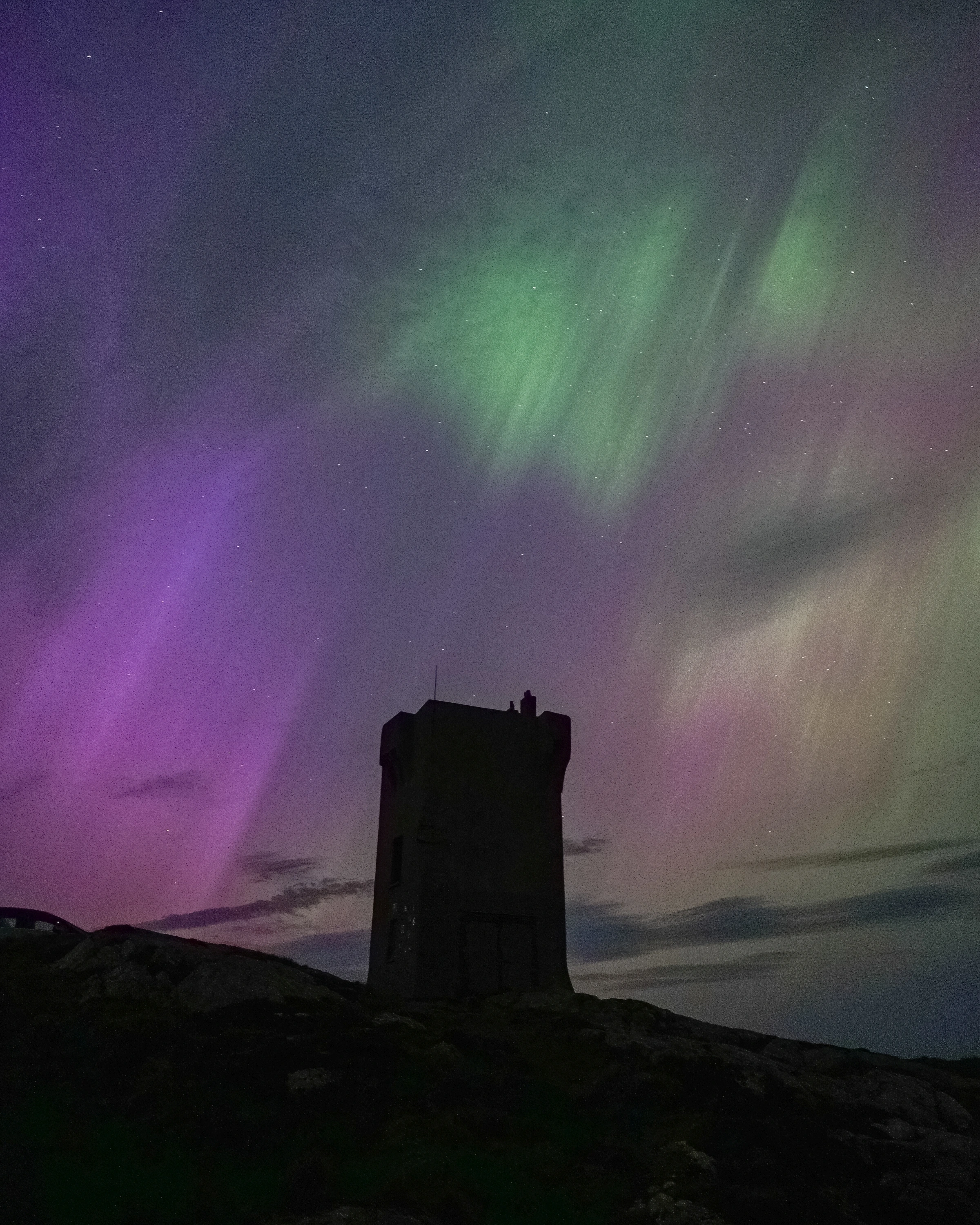 A dark stone tower and rocky ground sit beneath green and magenta aurora; part of a vehicle enters at the left.