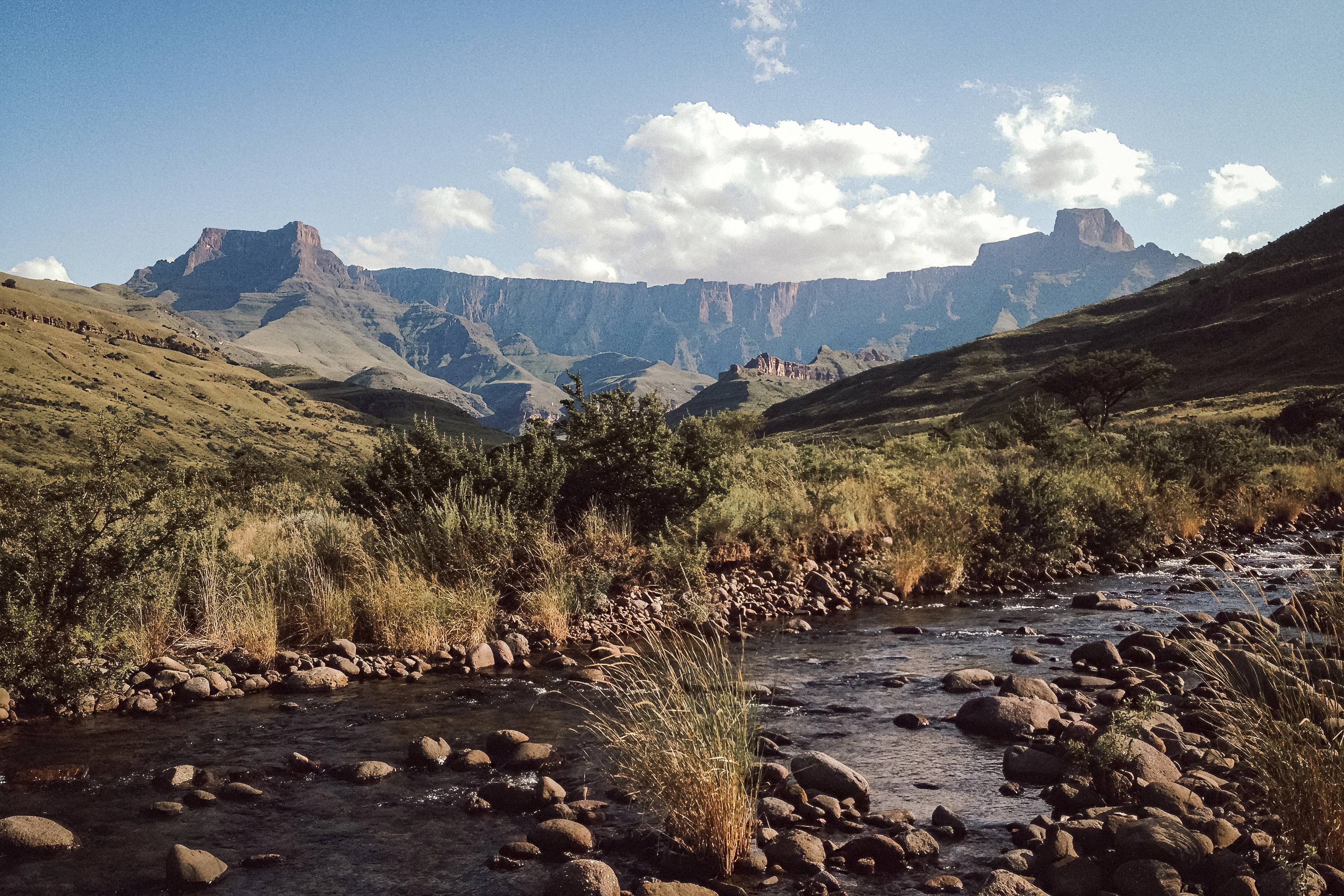 A rocky river and dry grasses lead toward a long mountain escarpment with prominent rock forms at either end.