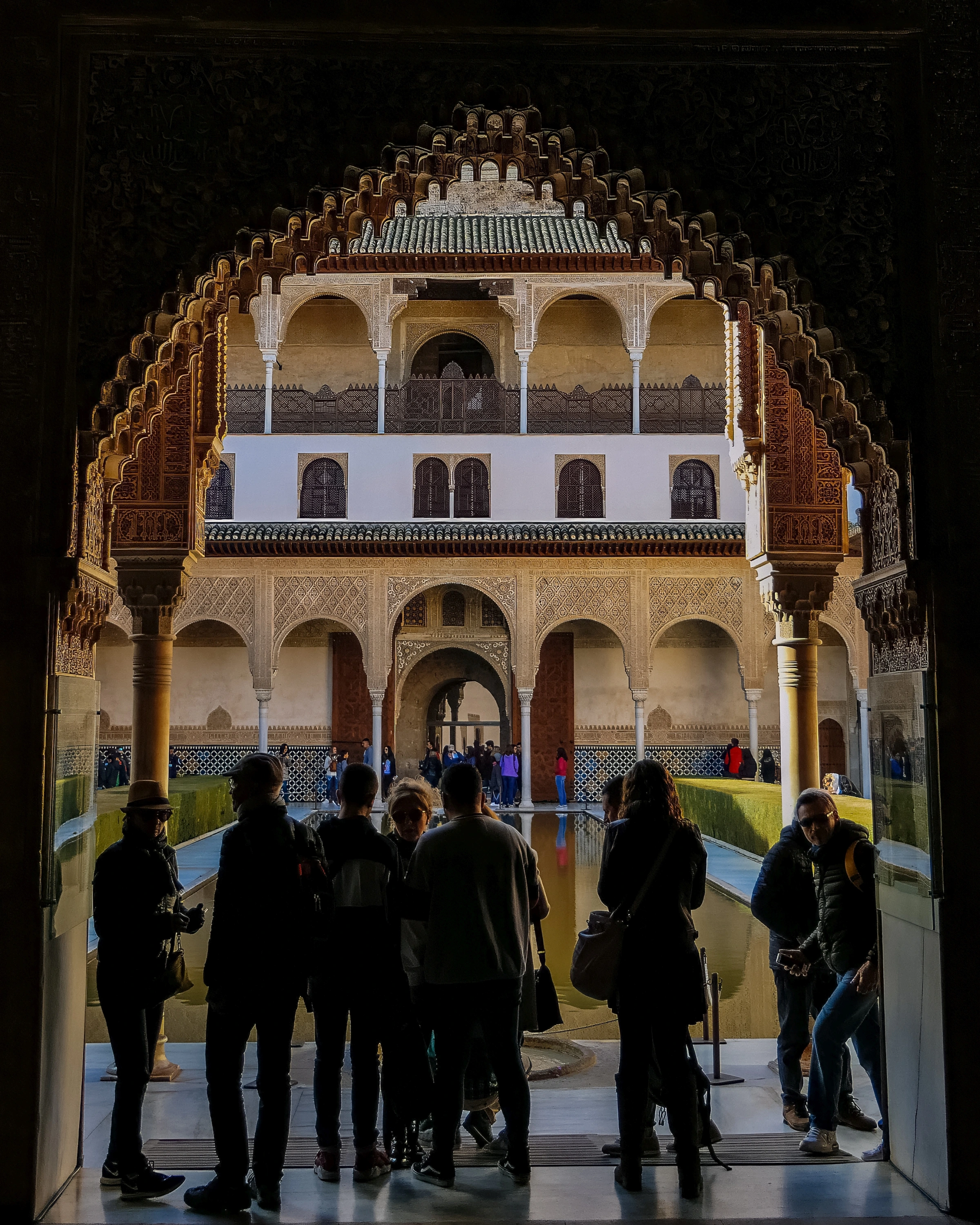 A dark ornate arch frames a bright courtyard, repeated smaller arches, water and several people.