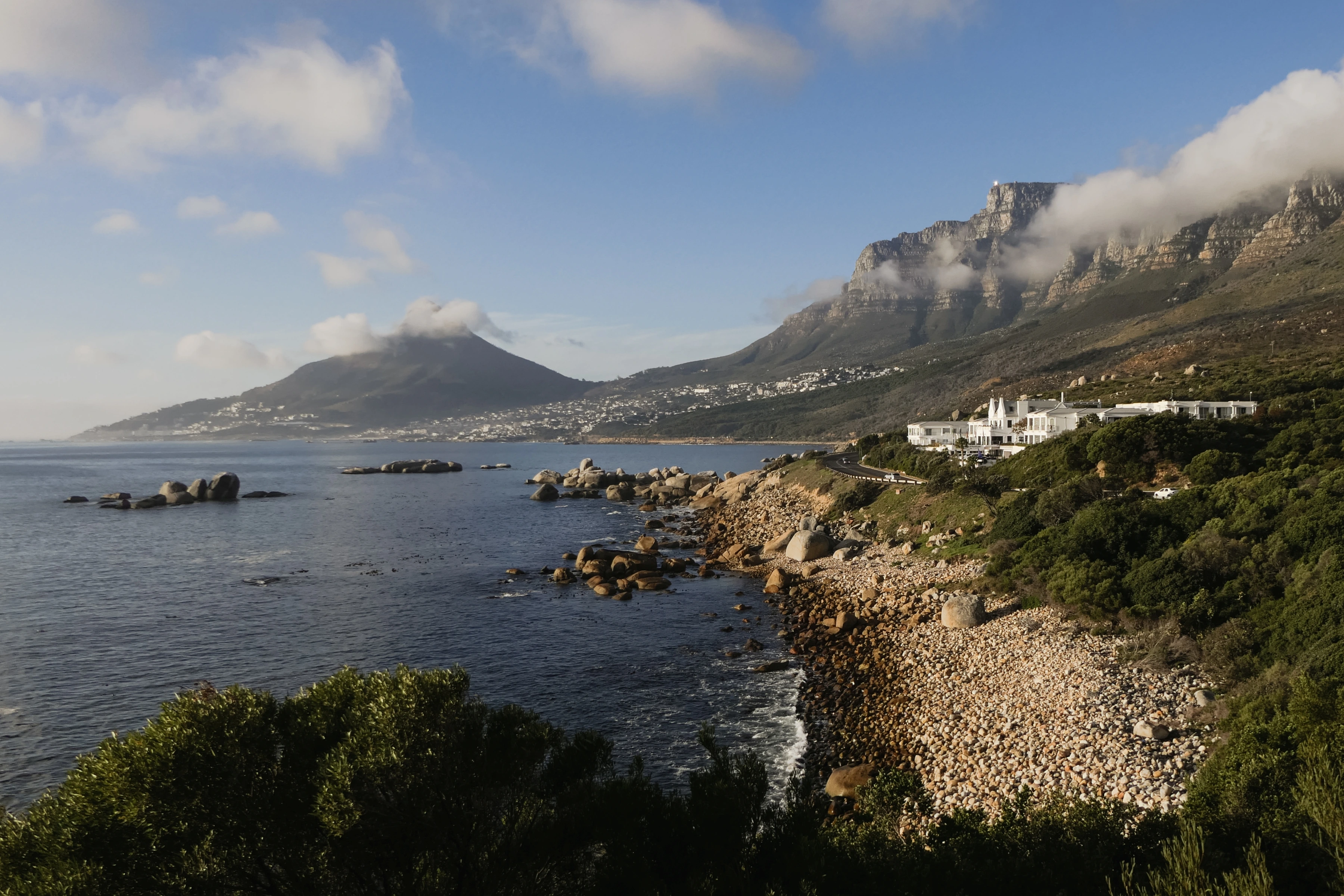 A road and white coastal buildings run beneath a cloud-covered mountain, beside boulders and the sea.