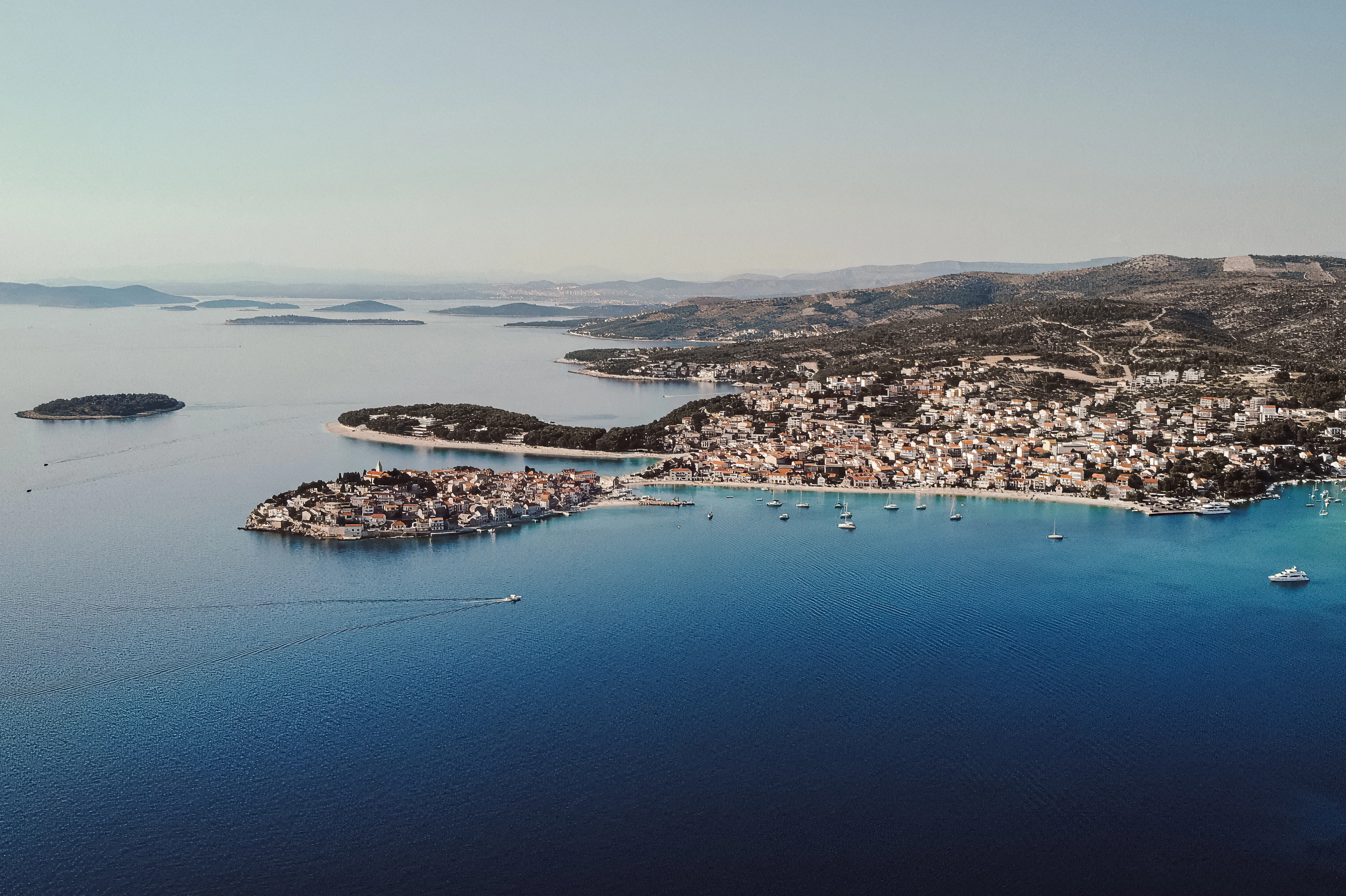 An aerial view follows a coastal settlement around a blue bay with a small boat wake.