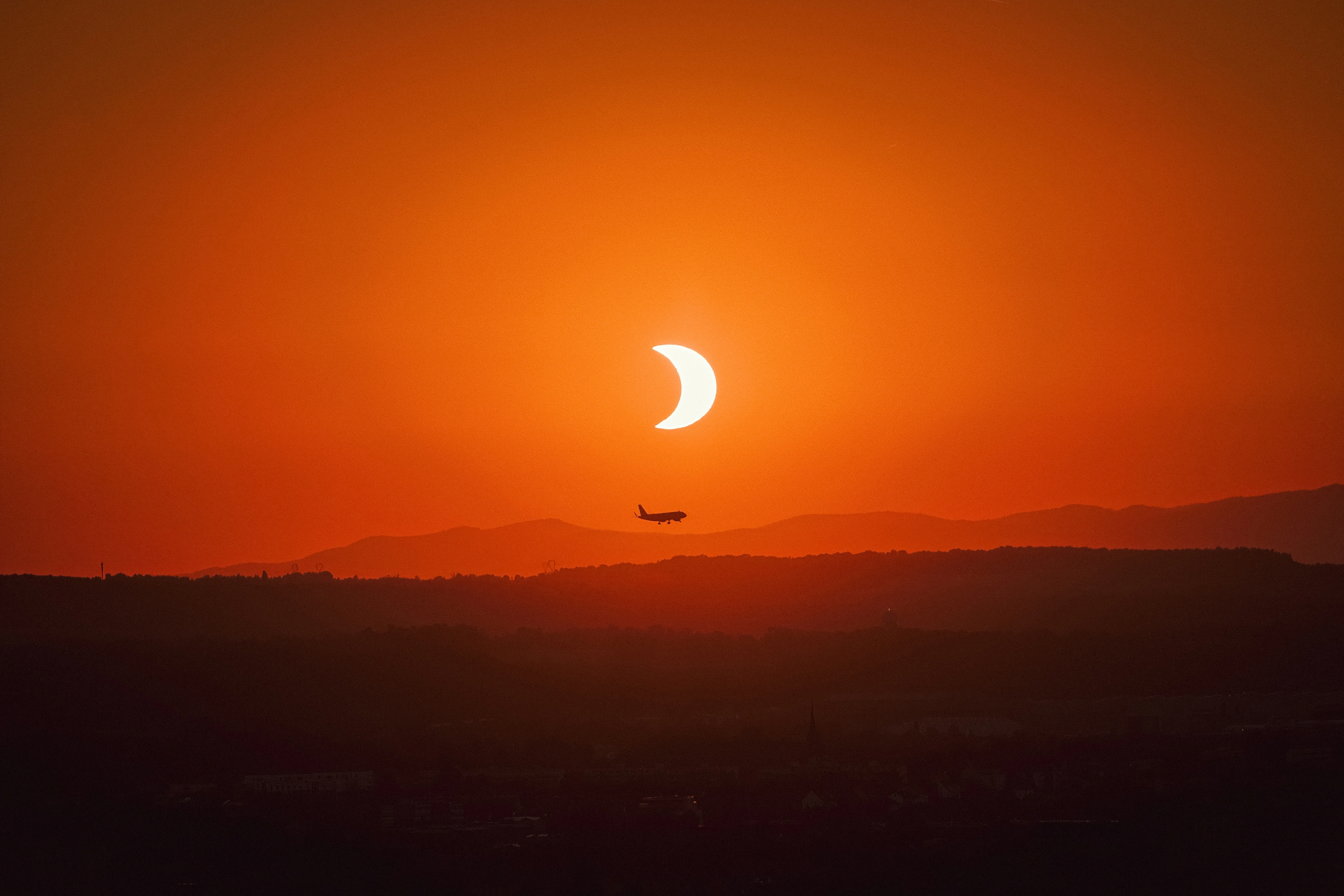 An aircraft passes beneath a bright solar crescent over warm layers of hills.