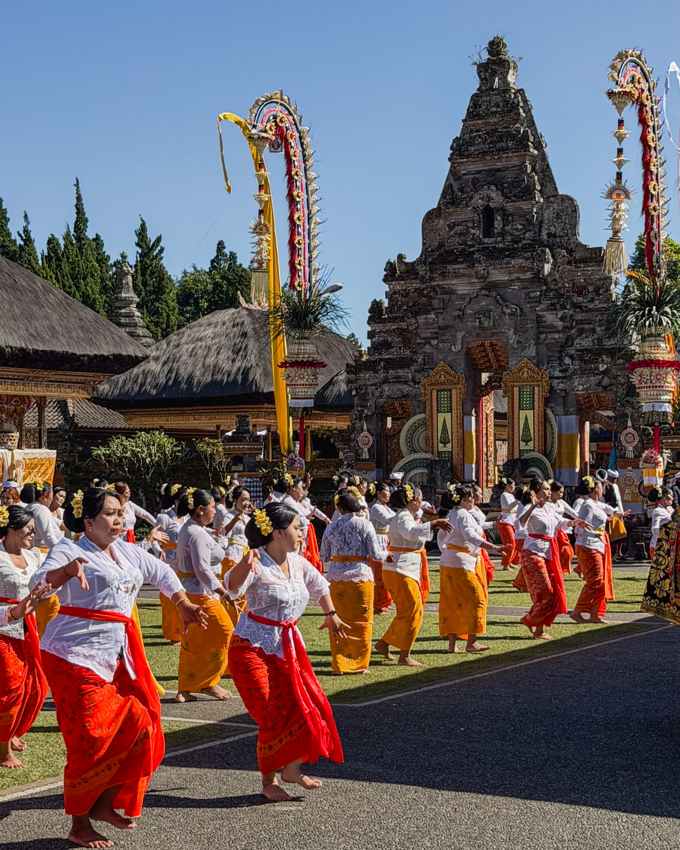 A group in white tops and bright red and yellow garments moves before a decorated stone gate.