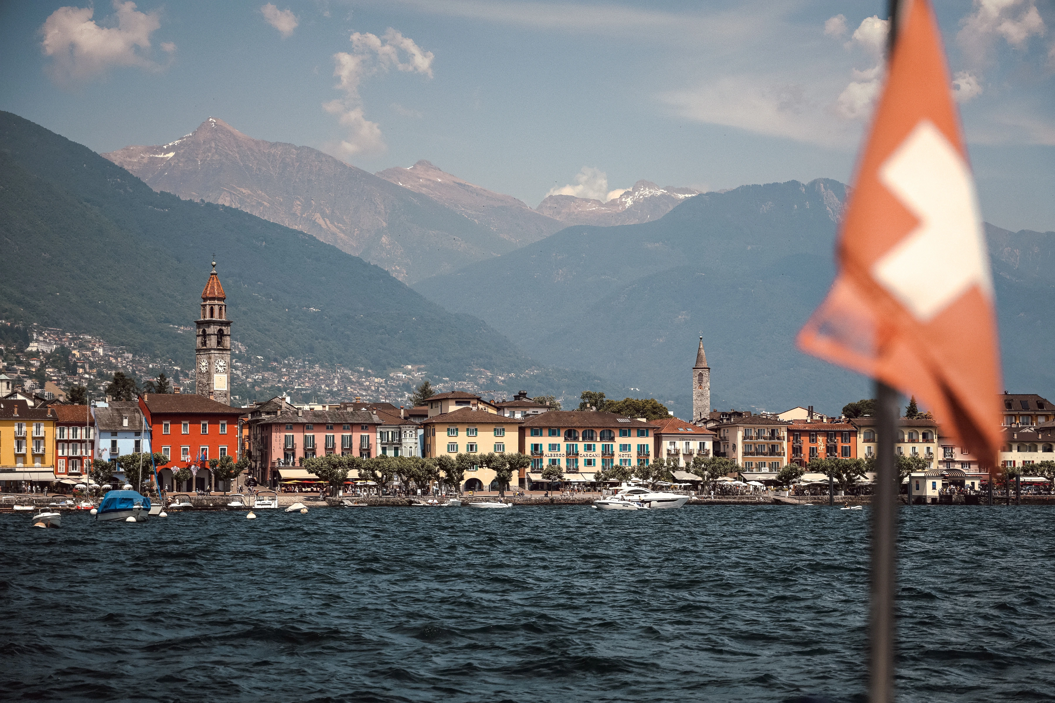 A soft Swiss flag occupies the right foreground before waterfront buildings, two towers and distant mountains.