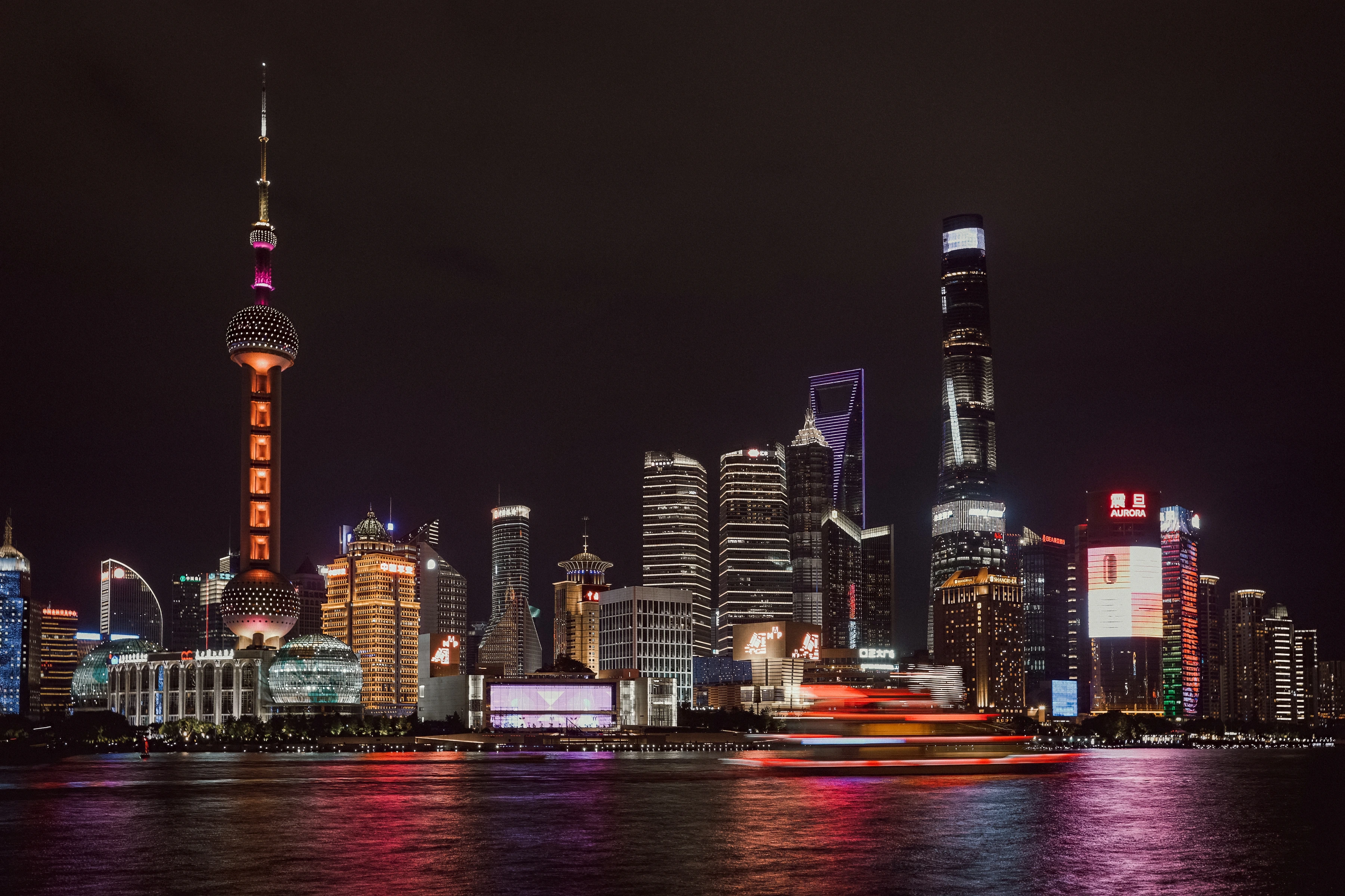 A moving red boat crosses below illuminated towers and coloured river reflections.