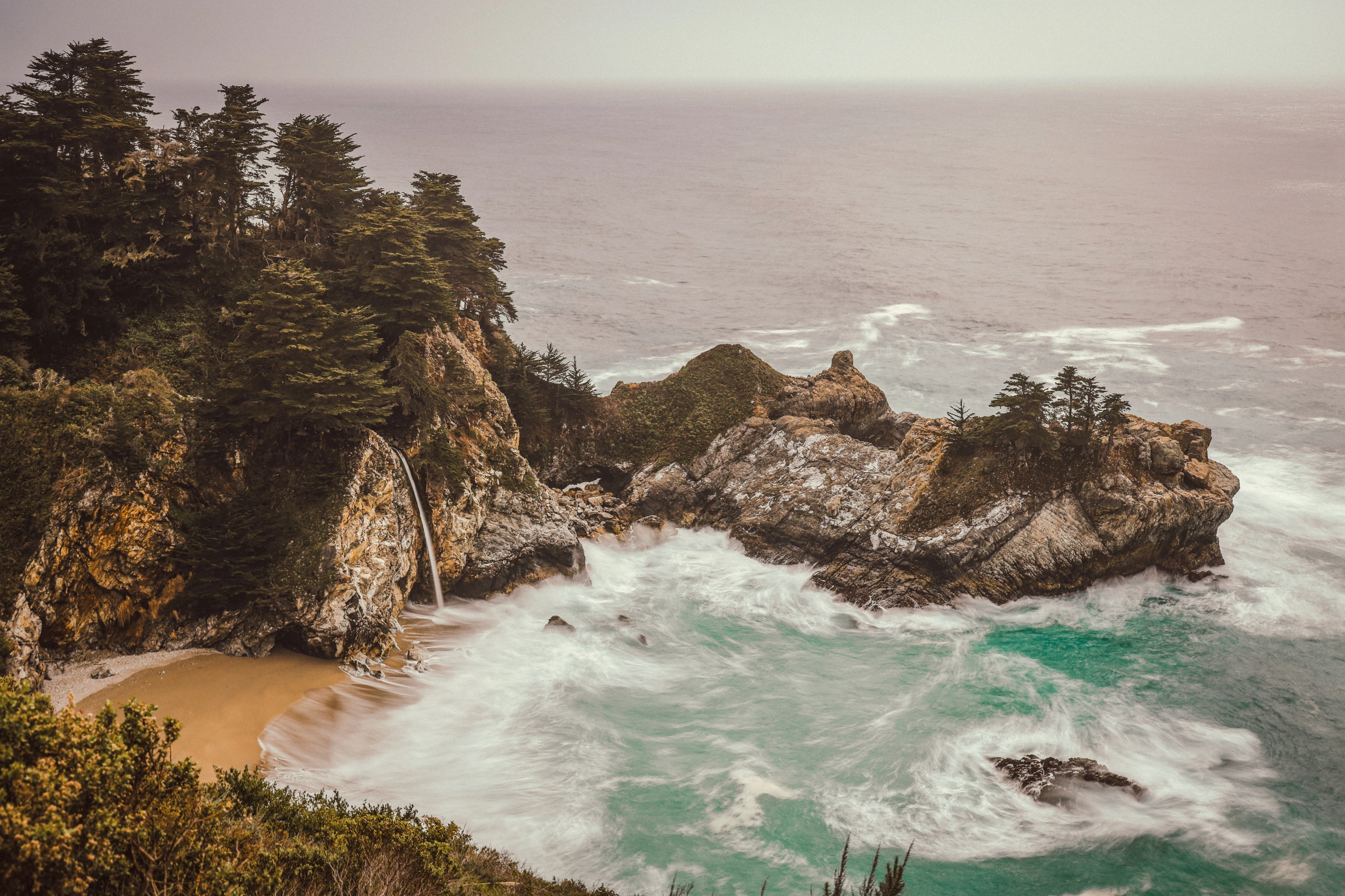 A narrow waterfall descends a wooded cliff beside a cove traced by white surf.