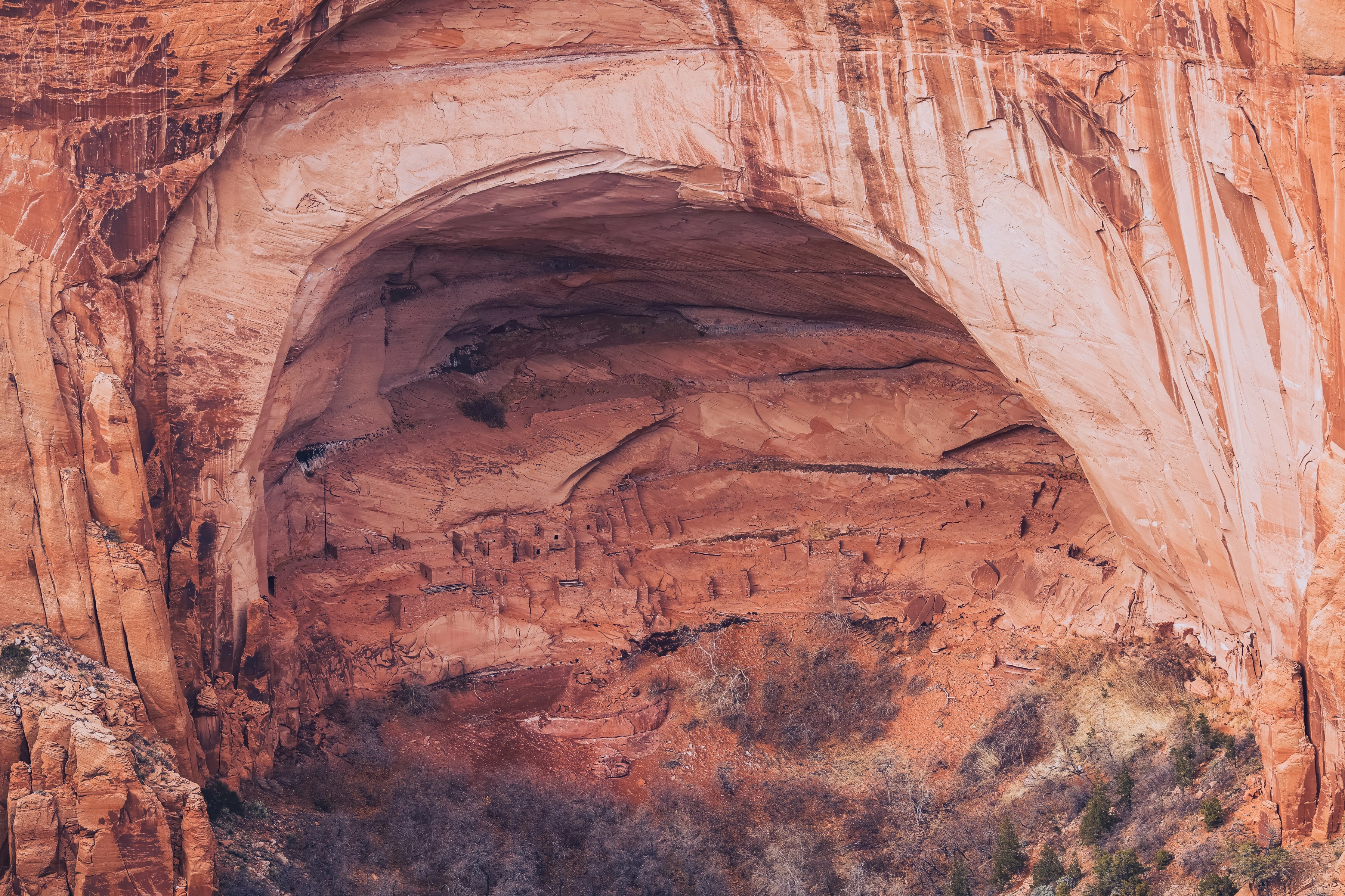 Small masonry rooms sit within a broad red rock alcove below a curving overhang.