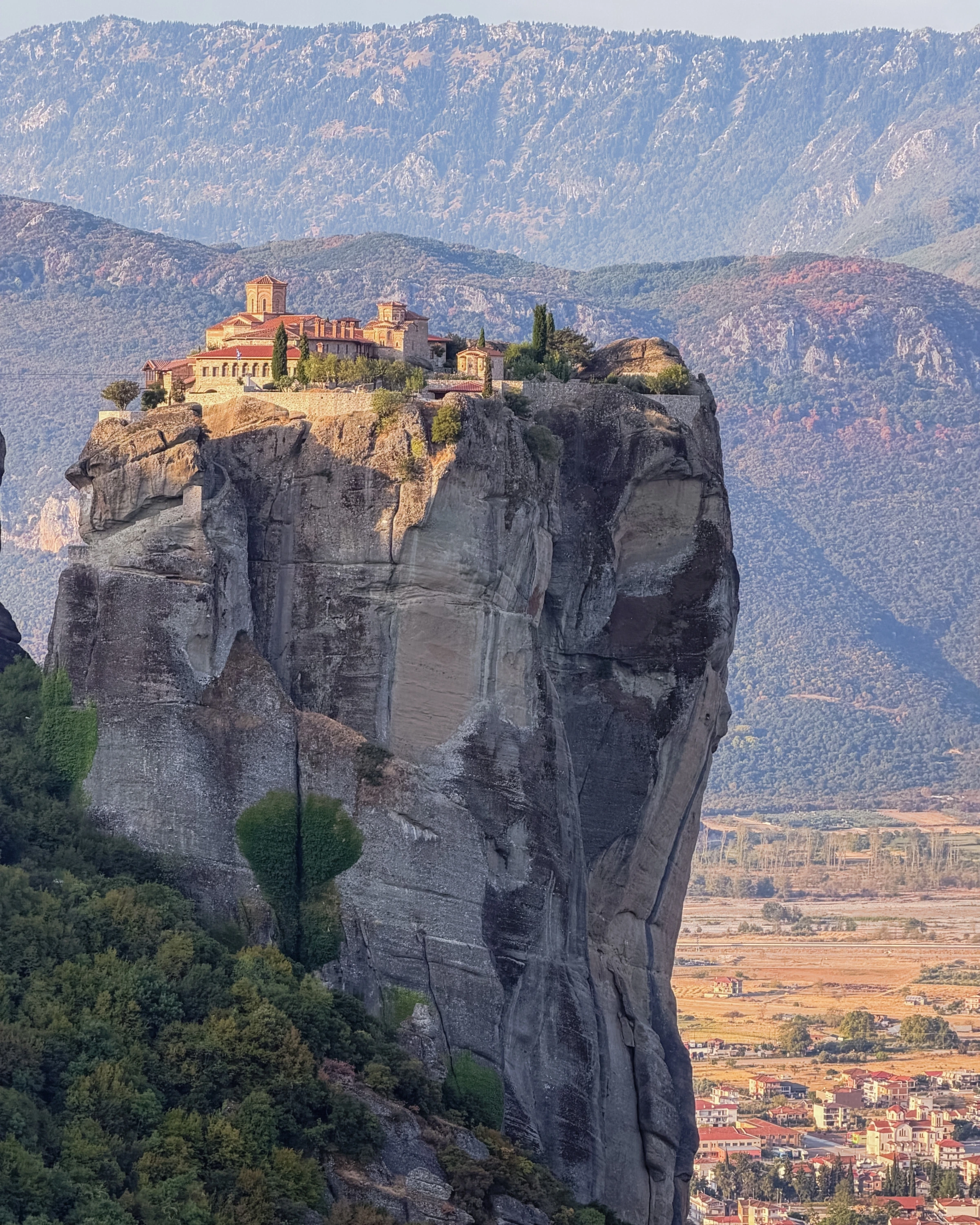 Buildings occupy the top of a tall rock pillar above a distant town and layered mountains.