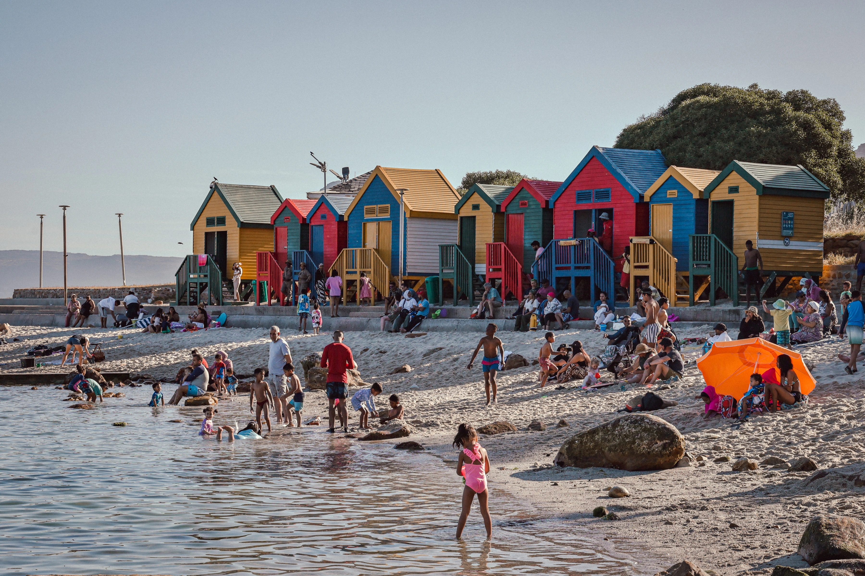 People spread along a beach in front of brightly coloured huts, with a child in the shallow foreground water.