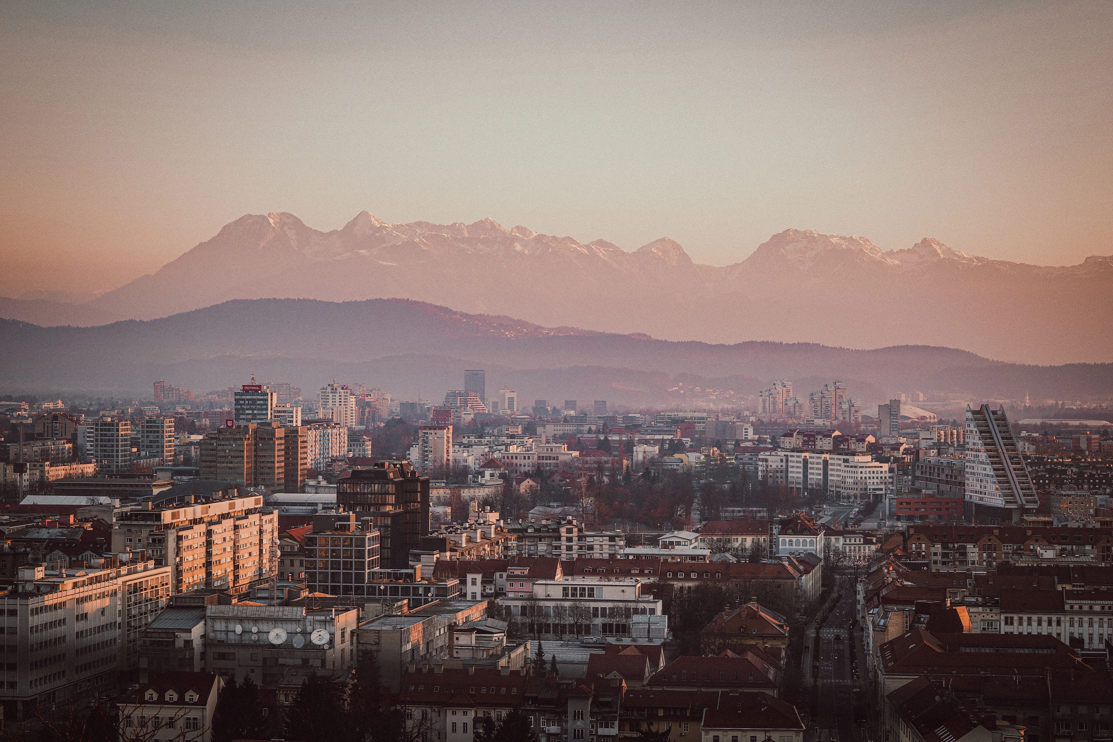 Modern towers and lower roofs sit beneath hazy foothills and pale snow-covered peaks in warm light.