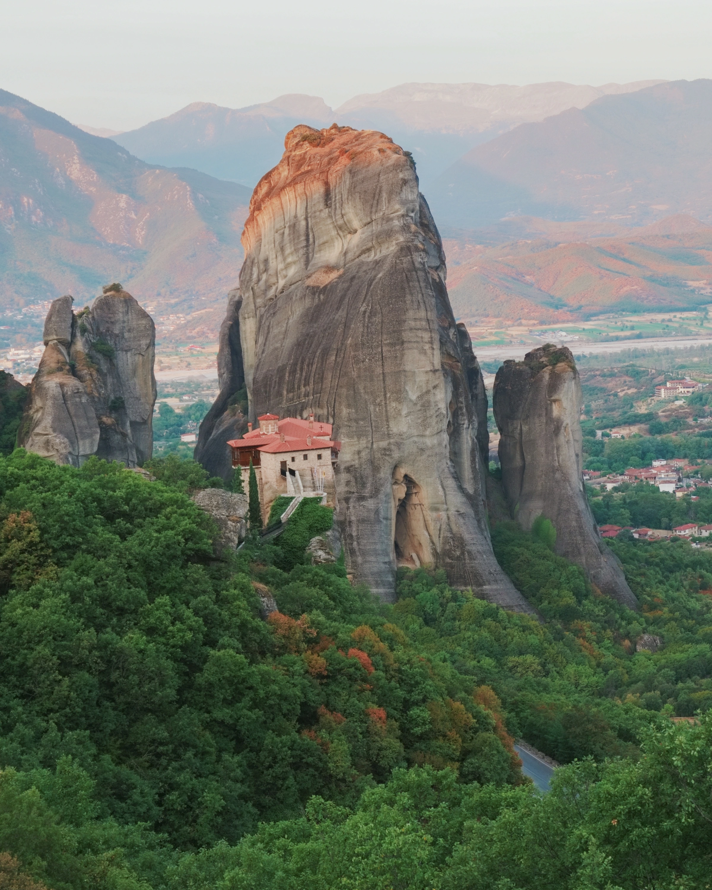 A red-roofed building sits below a tall rock pillar with wooded slopes and hazy mountains behind.