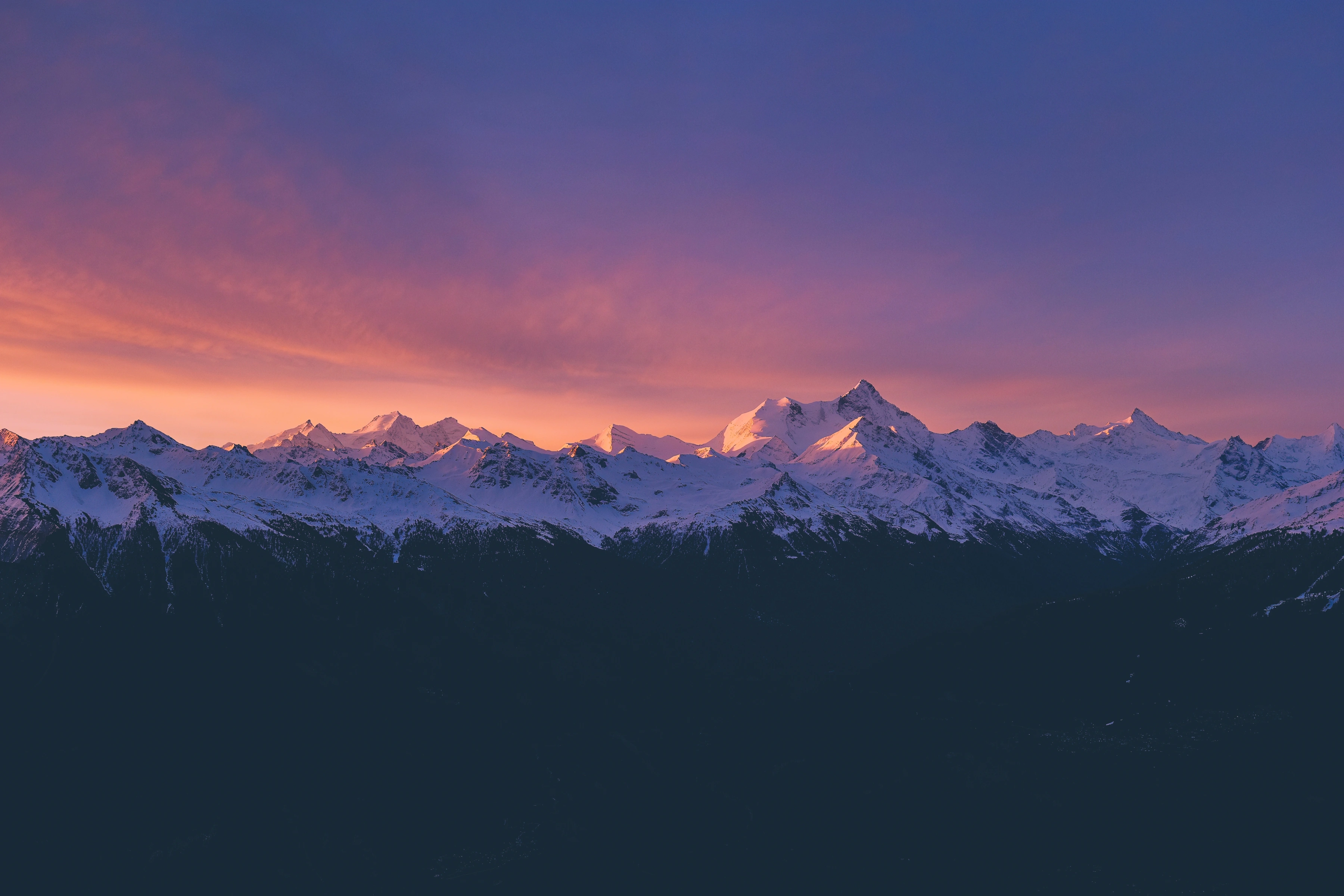 A snow-covered alpine ridge catches warm light below a pink and blue sky.