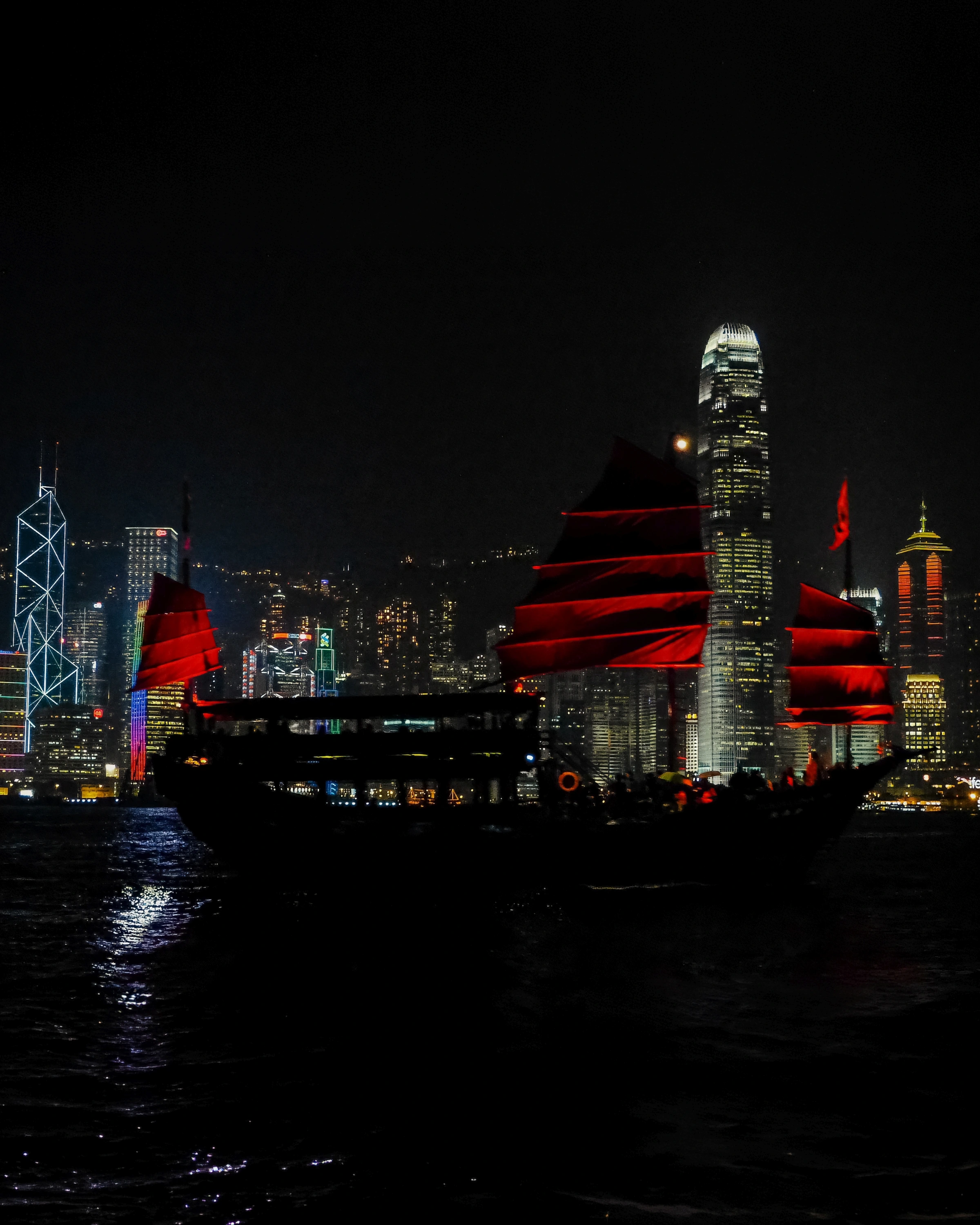 A red-sailed boat crosses a harbour in front of illuminated towers and their reflections.