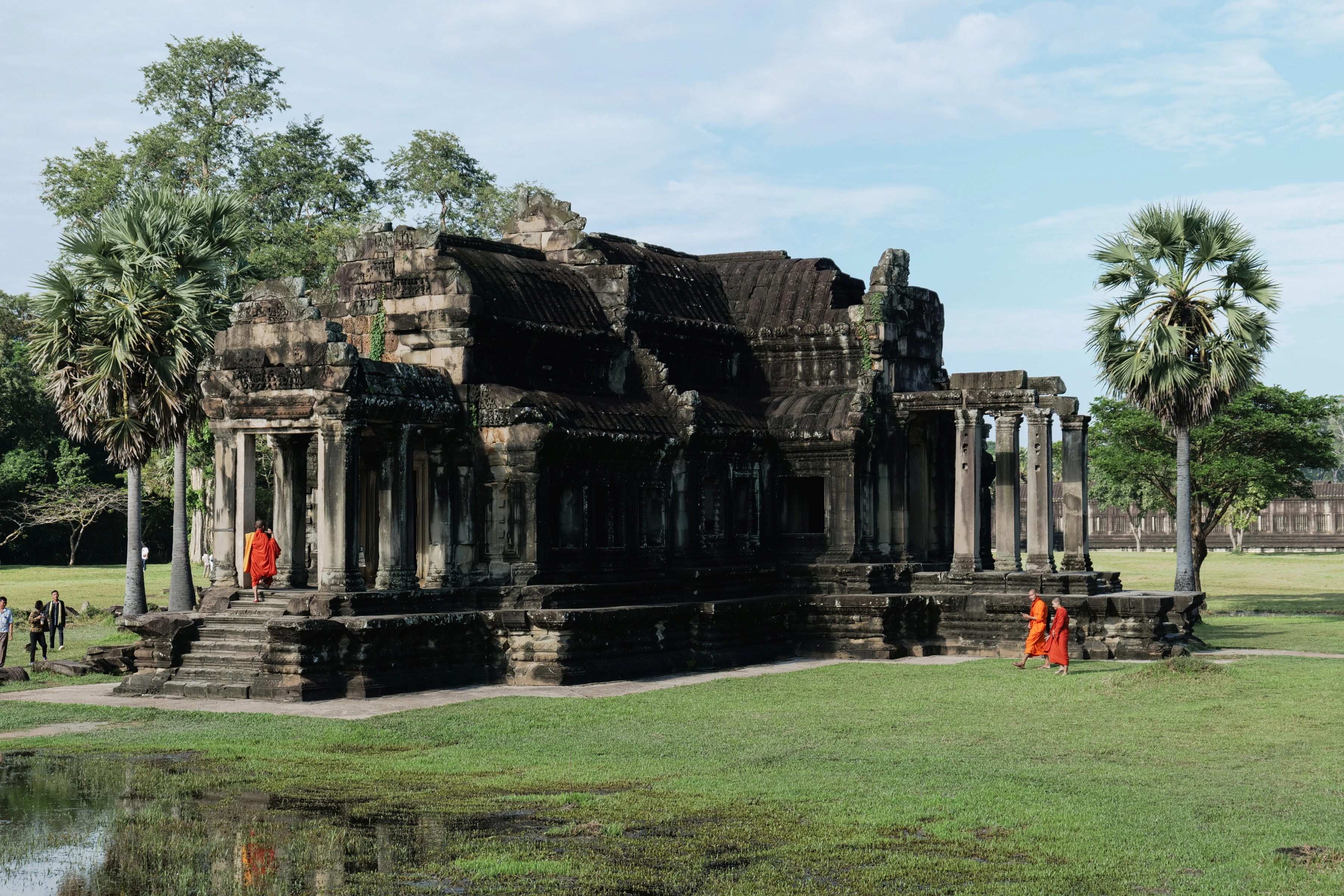 Orange-robed people appear around a stone pavilion between palms, with a small water reflection in the foreground.