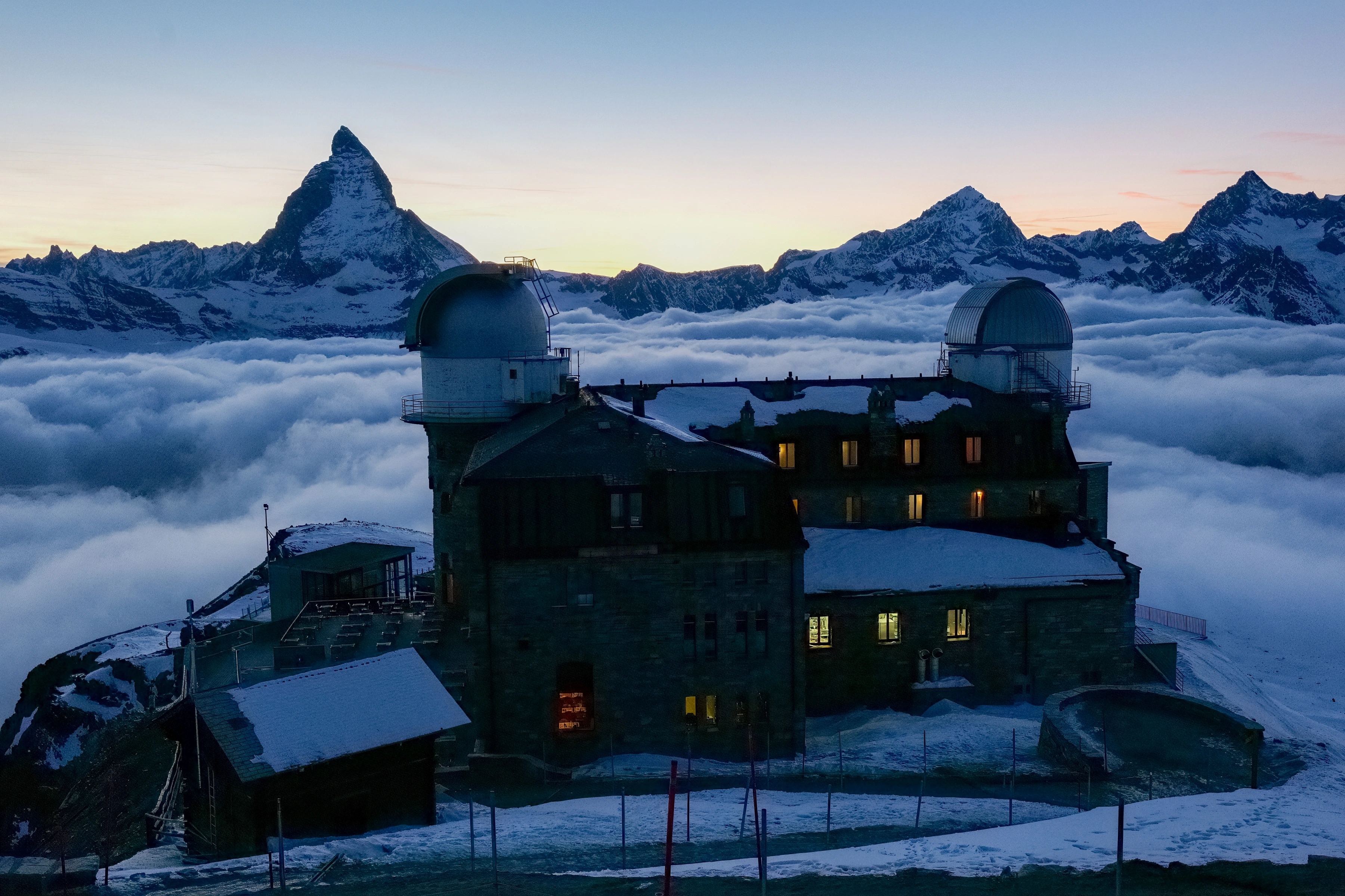 A dark mountain building with domes and warm windows stands above cloud, beside a sharp snowy peak and distant range.
