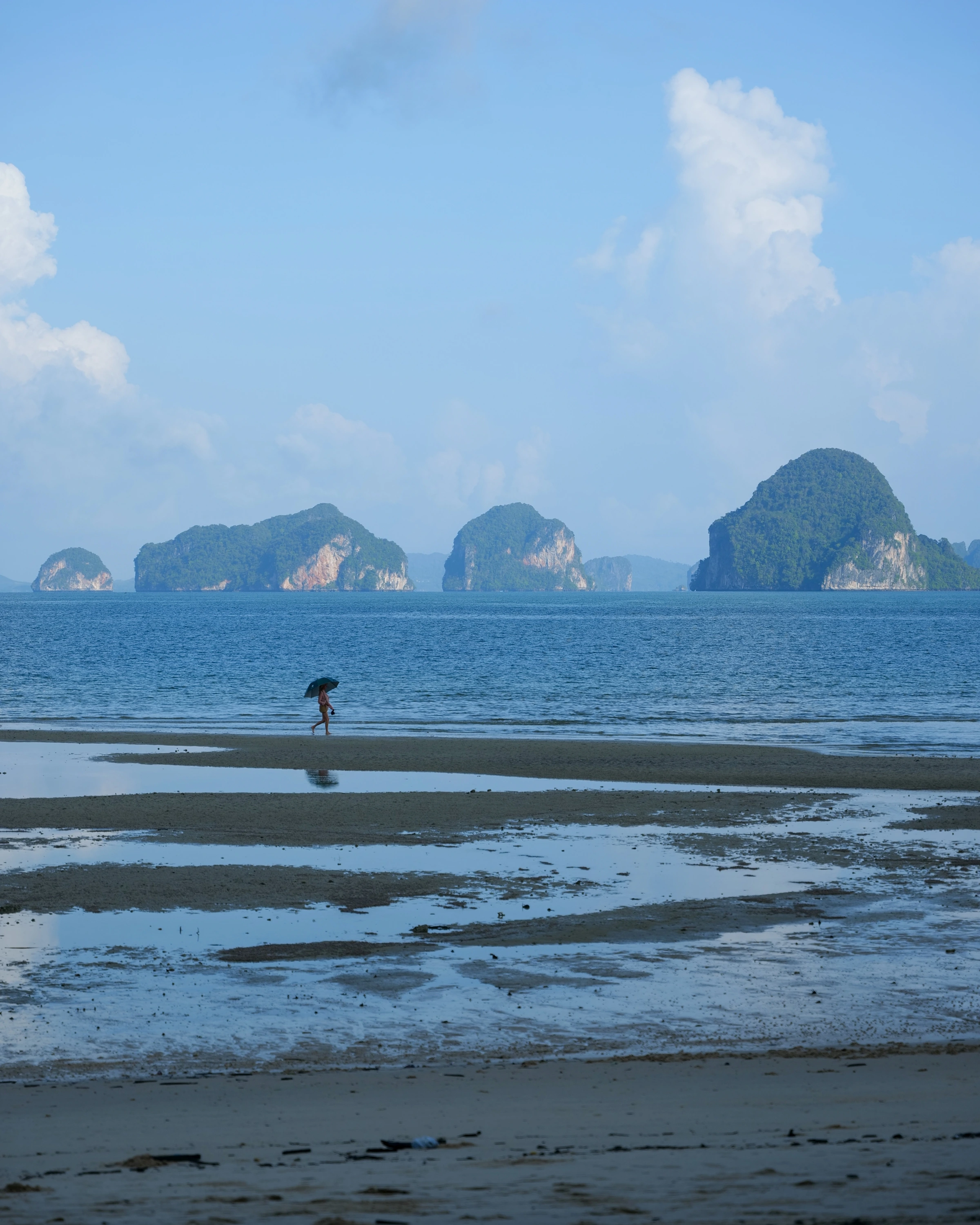 A person with an umbrella walks across a sandbar below rocky islands and pale sky.