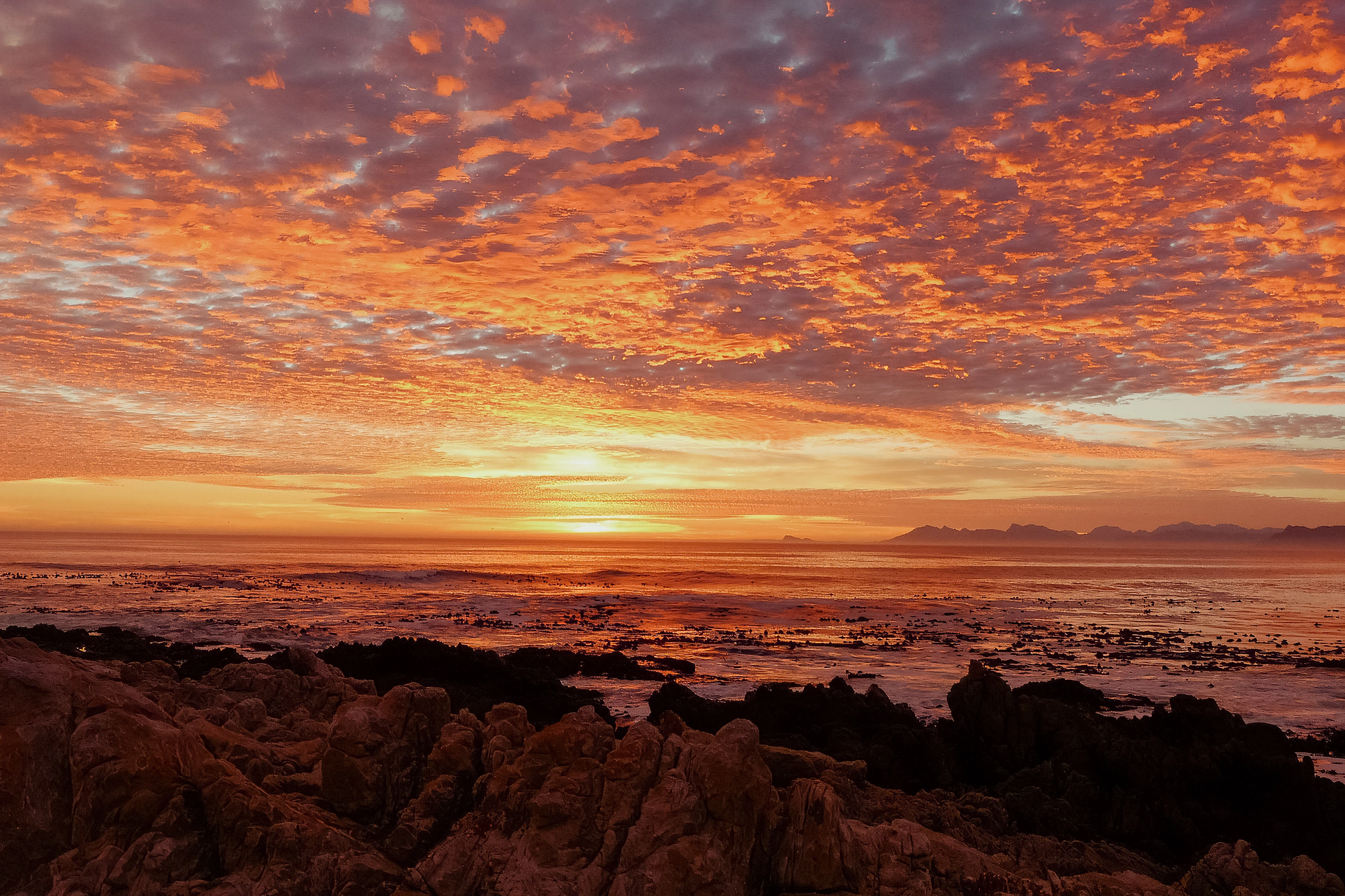Orange light spreads beneath layered cloud above a rocky shore, open water and a distant headland.
