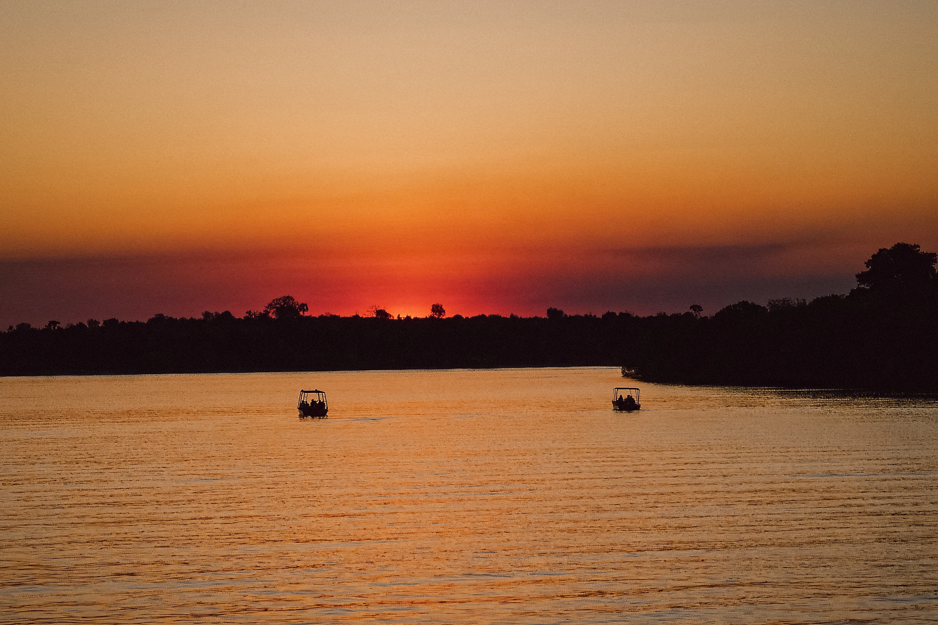 Two small boats sit on orange water beneath a dark wooded horizon.