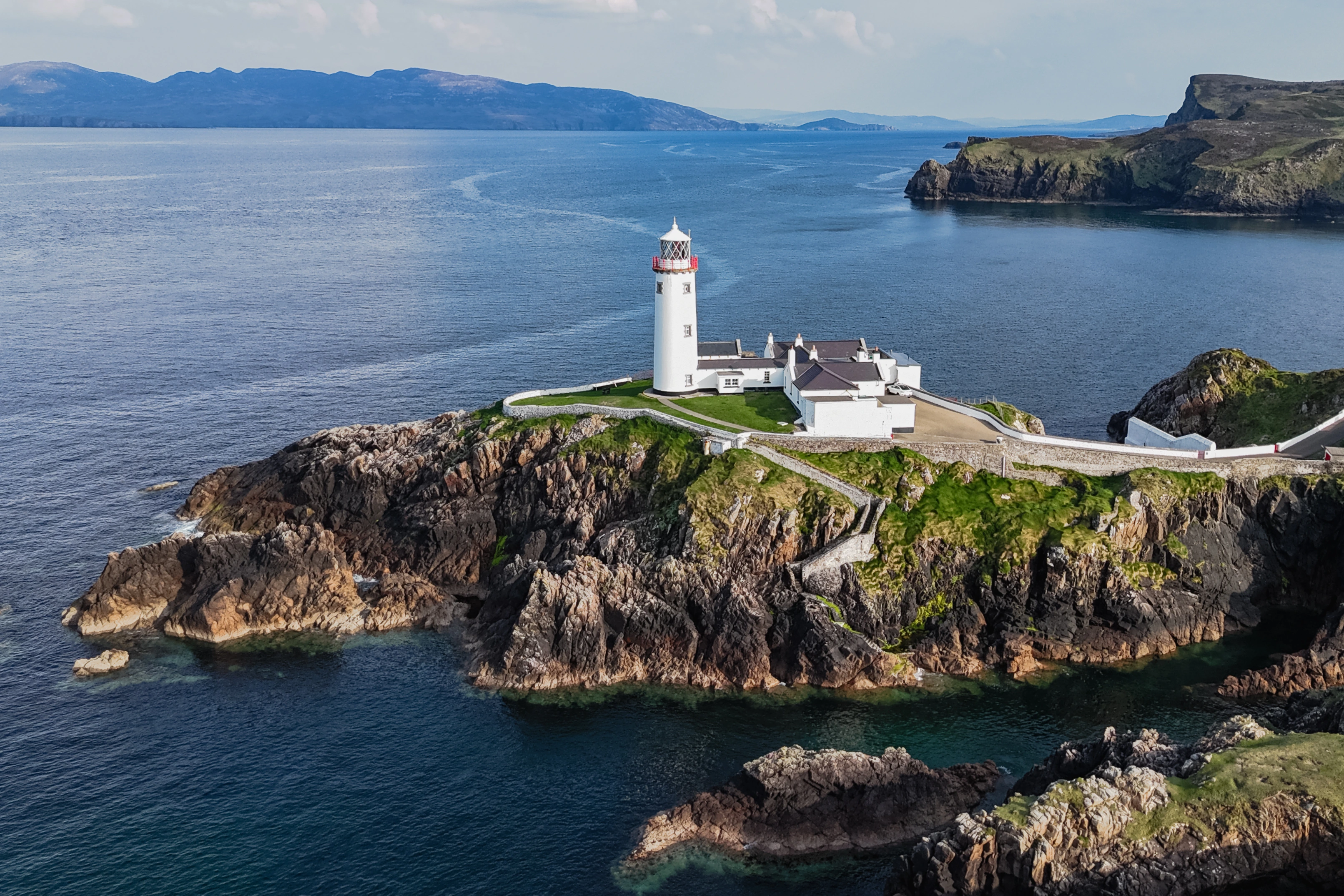 A white lighthouse with a red railing stands on a rocky coastal point.