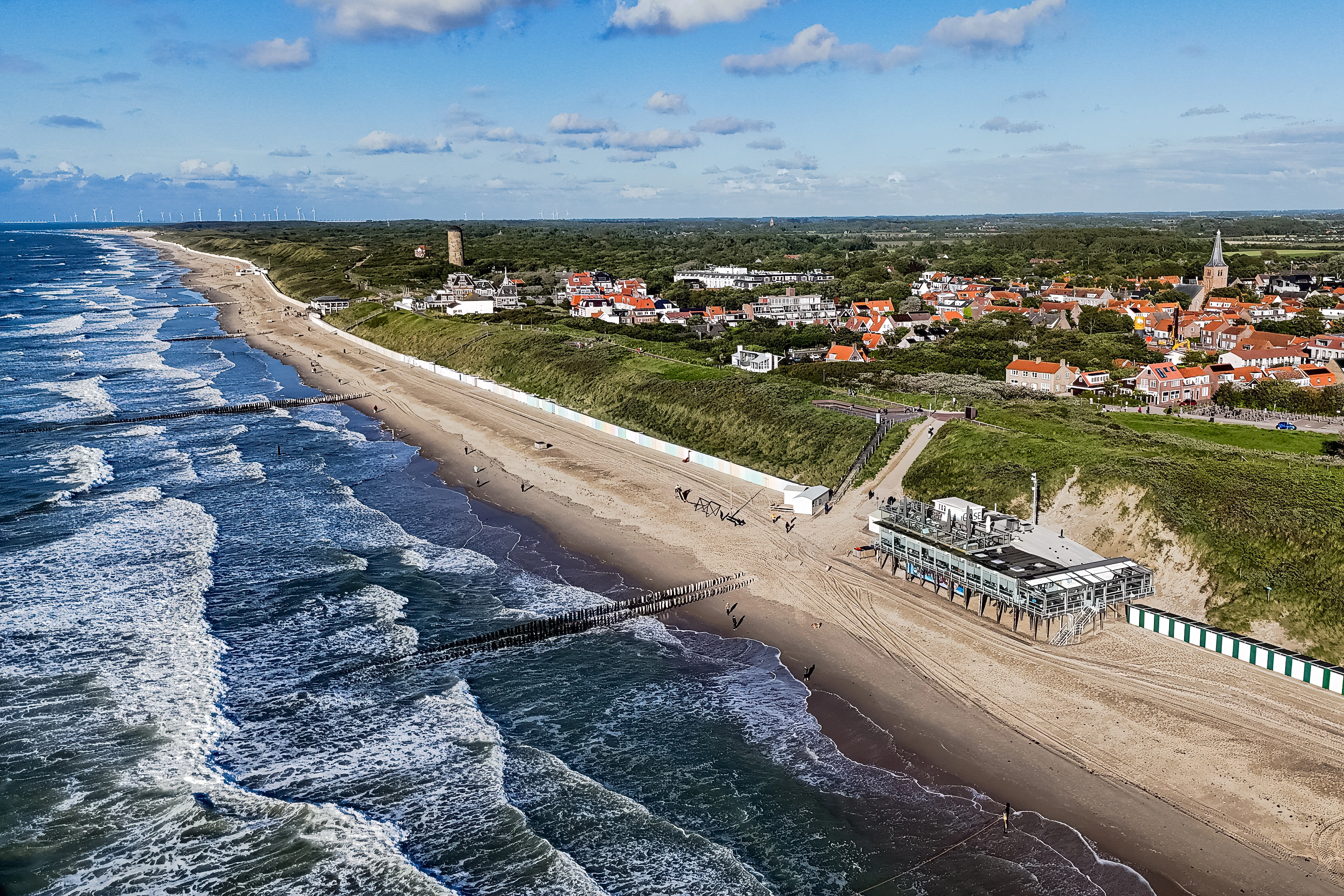 Parallel breakers and groynes repeat along a beach beside a coastal settlement.