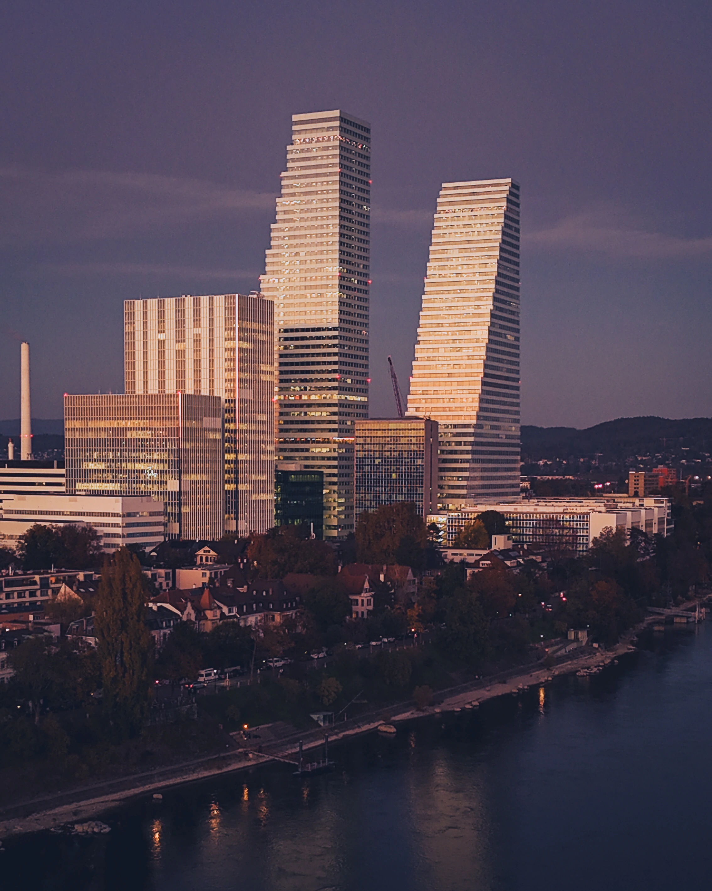 Two stepped modern towers and lower buildings catch warm light above a darker riverside neighbourhood.