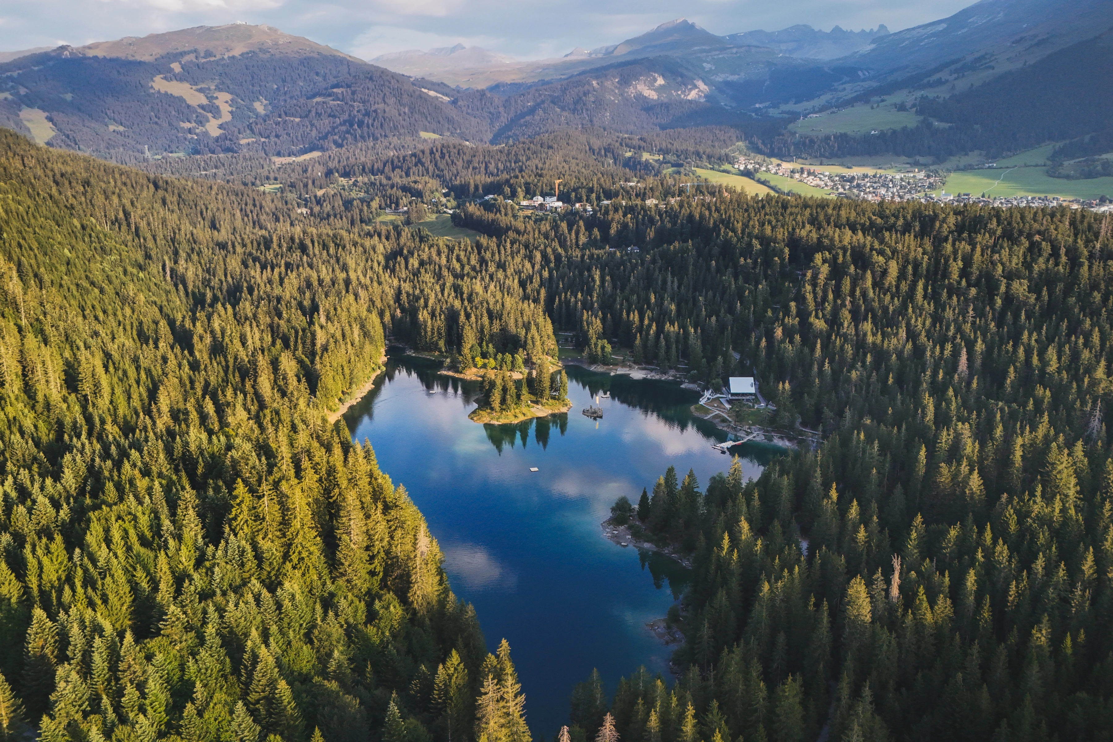 A wooded lake with a small tree-covered island sits below scattered buildings and mountain slopes.